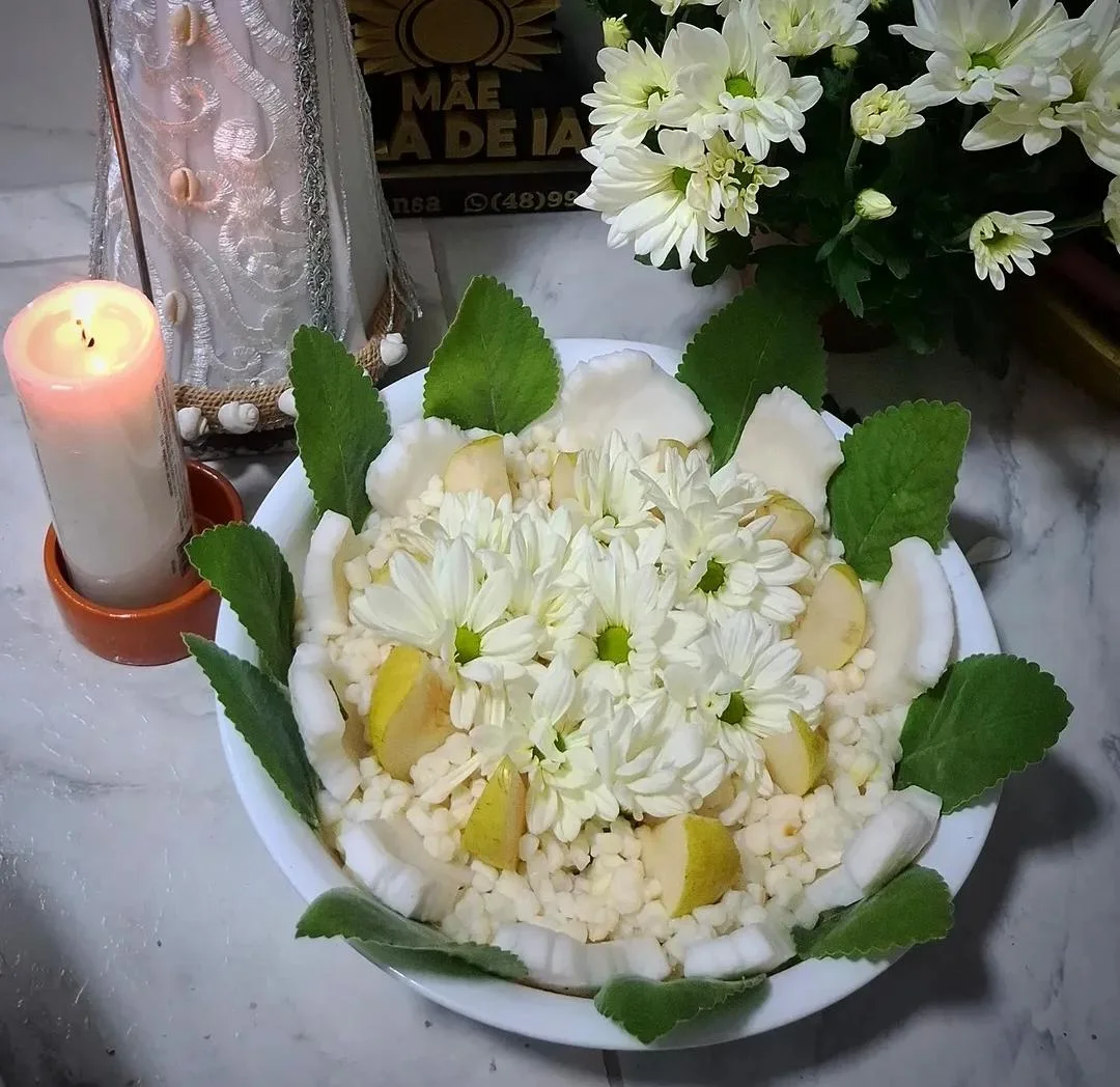 Oferenda branca em uma tigela com flores, frutas, grãos e folhas ao lado de uma vela acesa