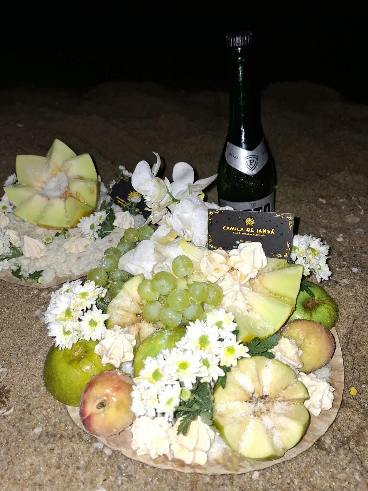 Oferenda sobre a areia com frutas verdes, flores brancas, doces e uma garrafa de espumante